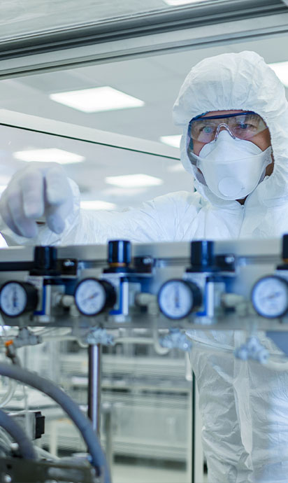 Ein Arbeiter in weißem Overall, Mundschutz, Schutzbrille und Handschuhen vor einer pharmazeutischen Produktionslinie.