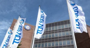 Drei Fahnen mit dem TÜV SÜD-Logo wehen vor einem modernen Bürogebäude mit dem TÜV SÜD-Schild an der Fassade.