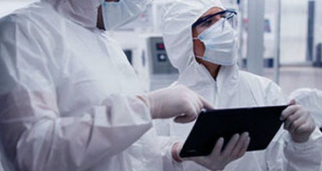 Zwei Arbeiter in weißen Overalls, Mundschutz, Schutzbrille und Plastikhandschuhen arbeiten in einer pharmazeutischen Fabrik auf einem Tablet.